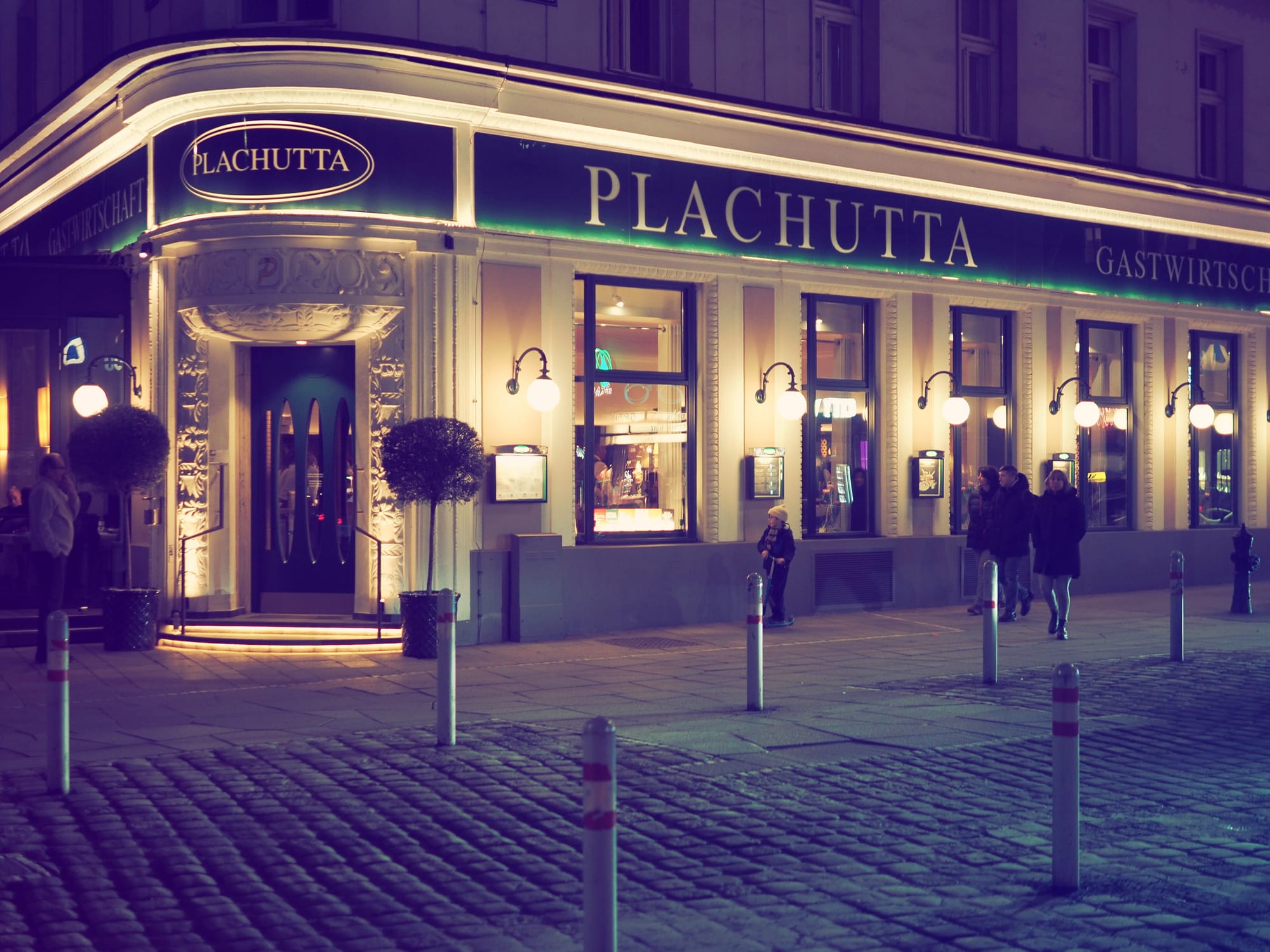 The illuminated Plachutta restaurant at night in Vienna, with pedestrians walking along the street and cobblestone pavement in the foreground.
