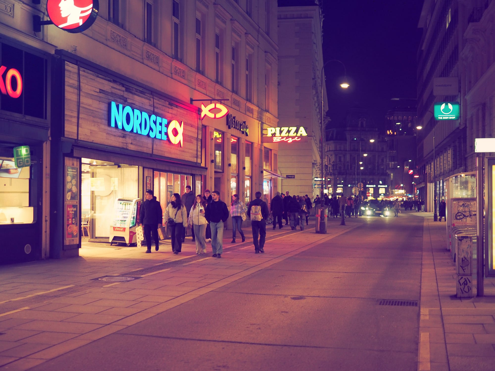 People walking along a busy street at night in Vienna, with illuminated shop signs and neon lights on both sides.